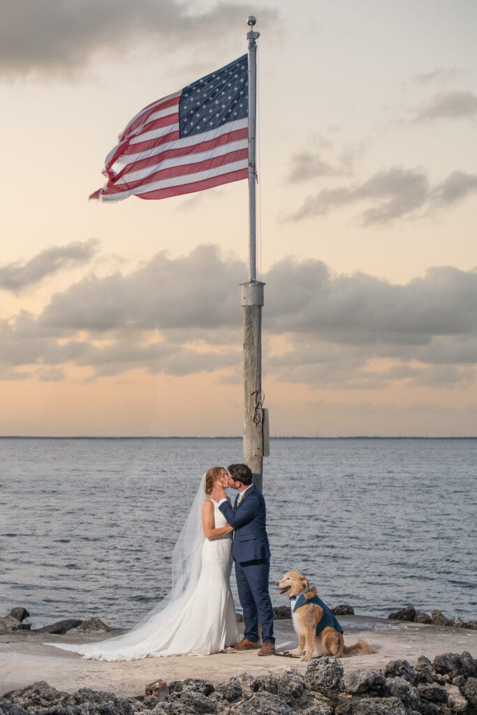 Florida Keys wedding couple kissing beneath American flag with their dog by the water