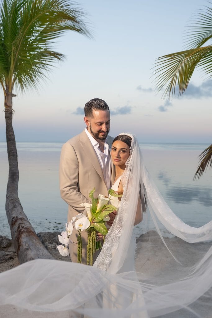 Bride and groom embracing by the waterfront with her veil flowing