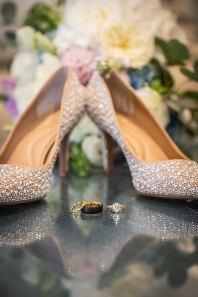 Sparkling bridal heels with wedding rings arranged on a reflective tabletop.