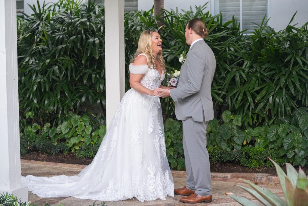 Amanda laughing and holding Trevor's hands during their first look at the Pelican Club, surrounded by tropical greenery.