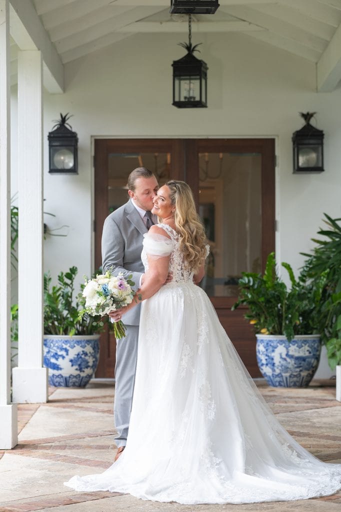 Trevor kissing Amanda's cheek as she holds her bouquet outside the Pelican Club, framed by tropical plants in blue and white pots. 