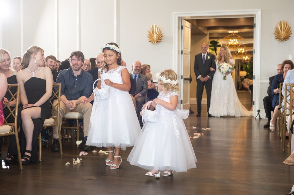 Two flower girls in white dresses and floral crowns carrying baskets down the ceremony aisle as guests watch, with Amanda waiting in the doorway behind them.