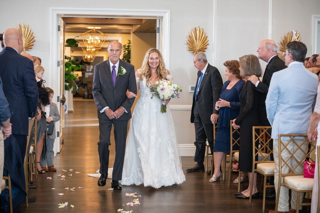 Amanda smiling as she walks arm in arm with her father down the petal lined aisle at the Pelican Club