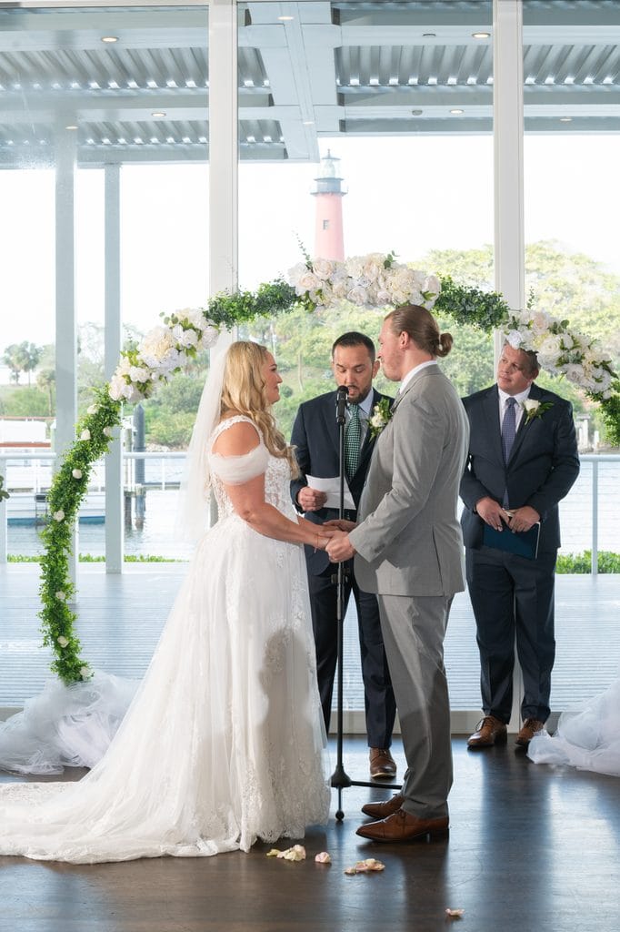 Amanda and Trevor holding hands beneath a white floral arch during their ceremony, with the Jupiter Lighthouse visible through the windows.