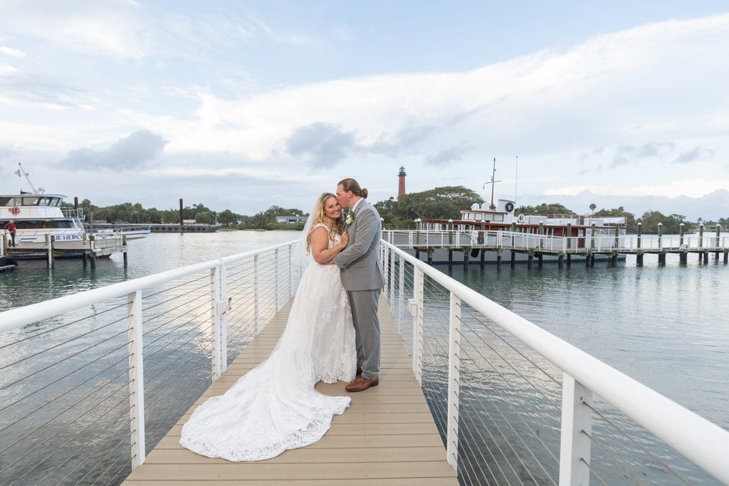 Trevor kissing Amanda's forehead on the Pelican Club dock, with her wedding dress train spread behind her and the Jupiter Lighthouse in the distance. 