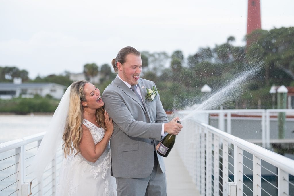Trevor spraying champagne as Amanda laughs beside him on the Pelican Club waterfront walkway. 
