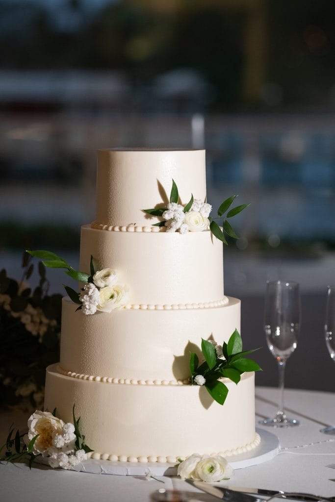 Four tier ivory wedding cake decorated with white flowers, greenery, and pearl style borders beside champagne flutes.