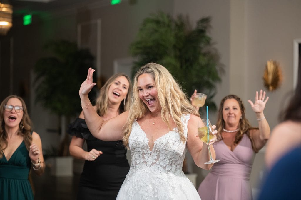 Amanda cheering with one hand raised and a drink in the other while dancing with guests at the reception.