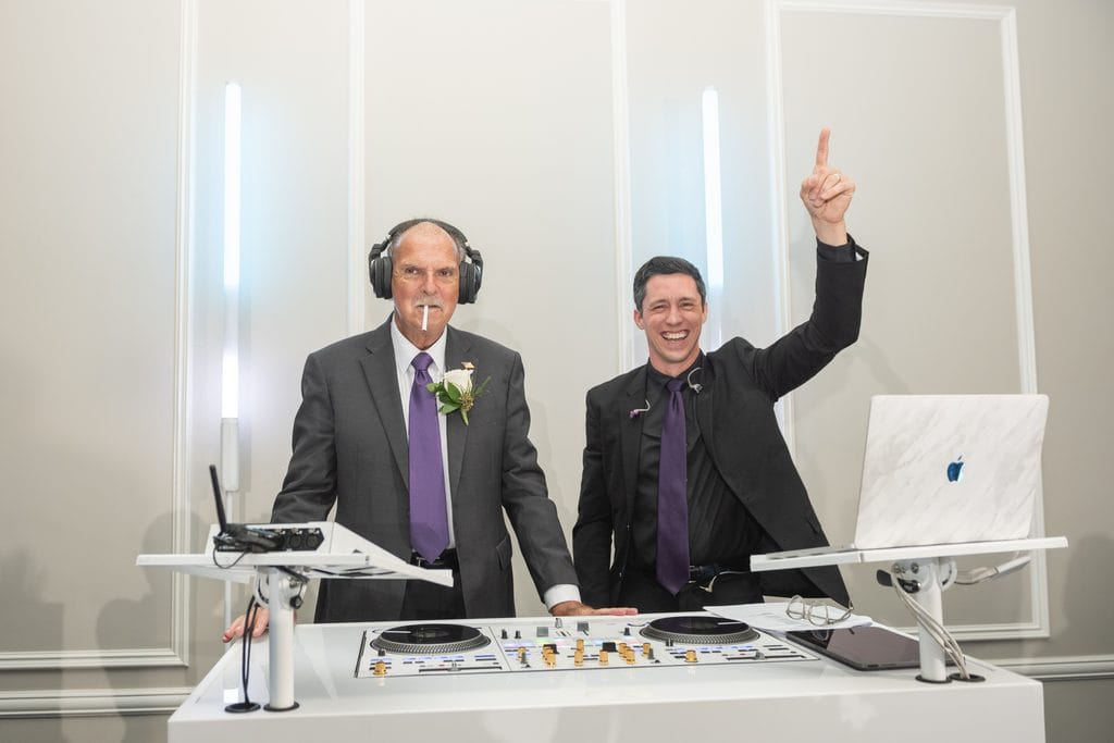 A wedding guest wearing headphones besides the DJ, who smiles and raises one finger behind the reception DJ booth.